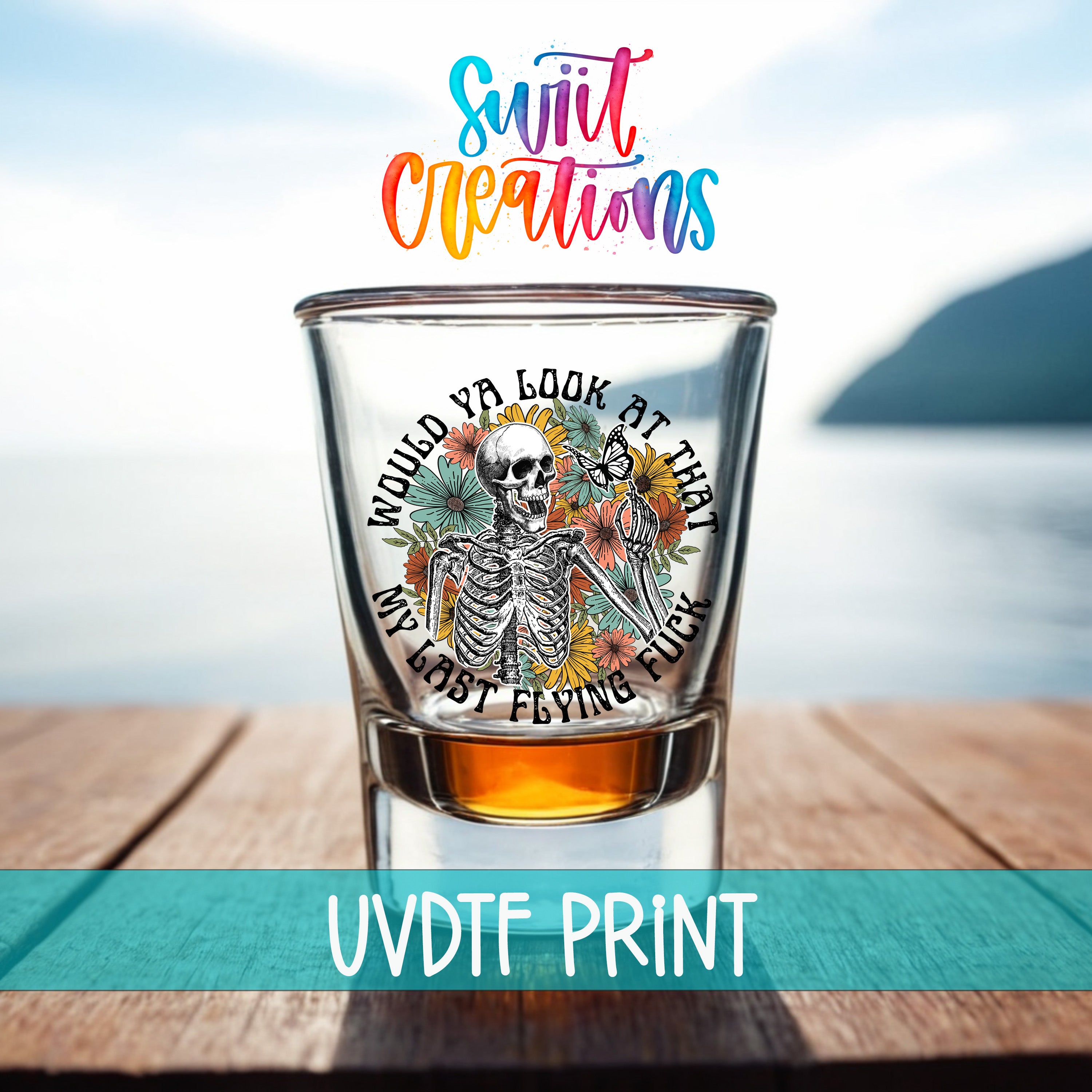 A shot glass with a skeleton design and text printed on it, placed on a wooden surface with a body of water in the background.