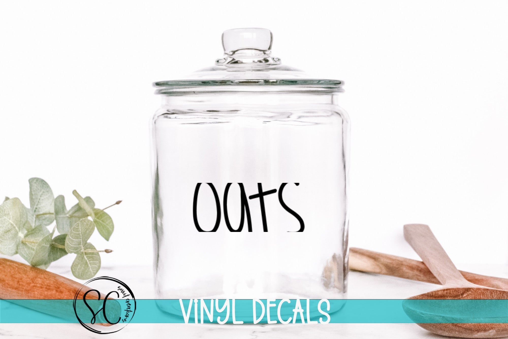 A clear glass jar with the word "Oats" printed on it, placed on a white surface next to a wooden spoon and some green leaves.