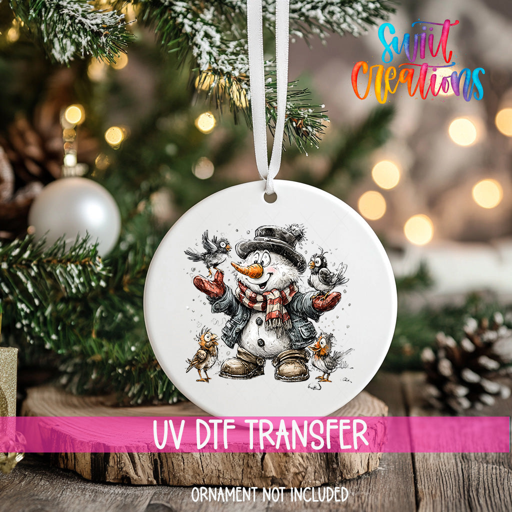 A white ornament with a snowman and birds is hanging on a Christmas tree, with a pink ribbon around it.