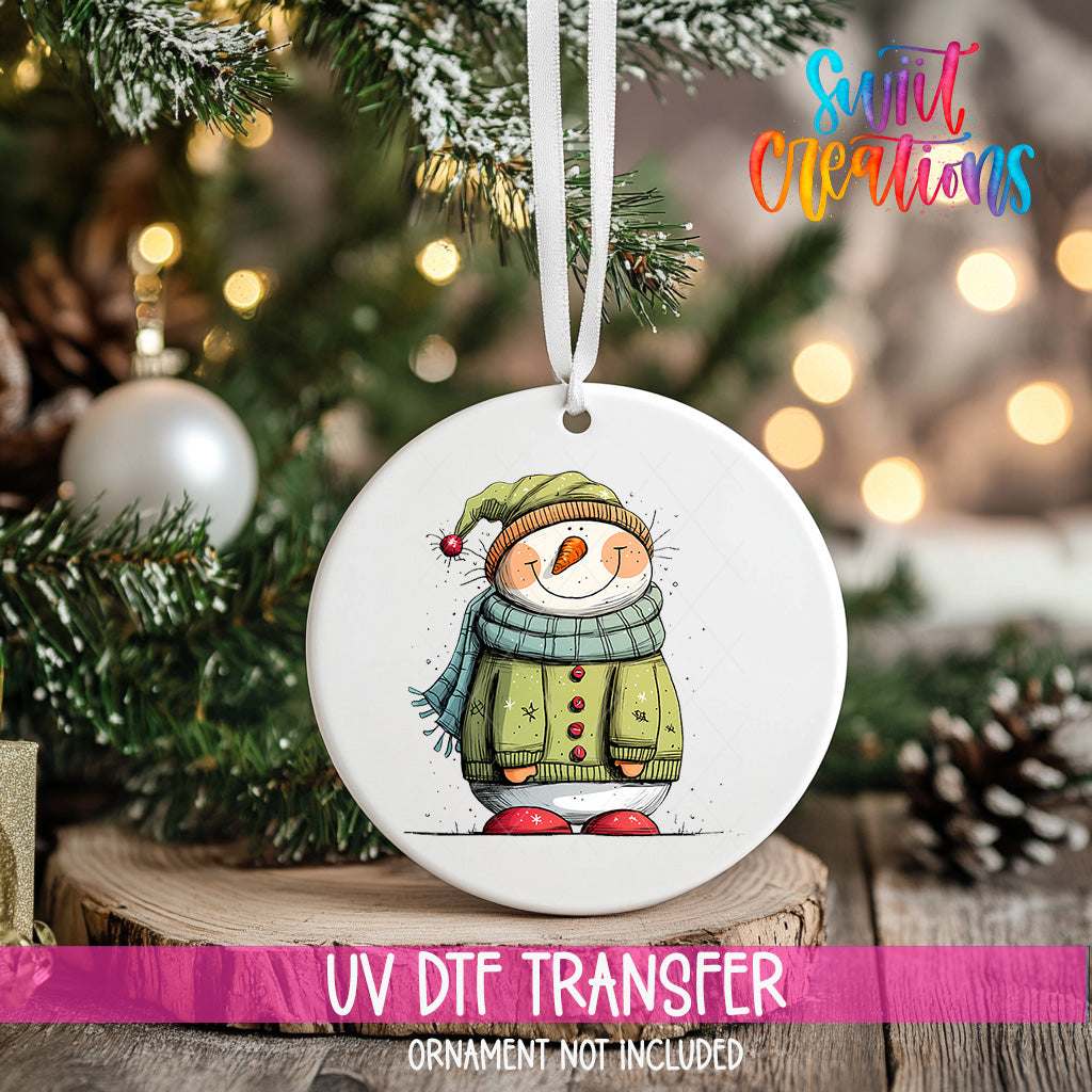 A white ornament with a snowman design hangs on a Christmas tree, surrounded by pine branches and twinkling lights.