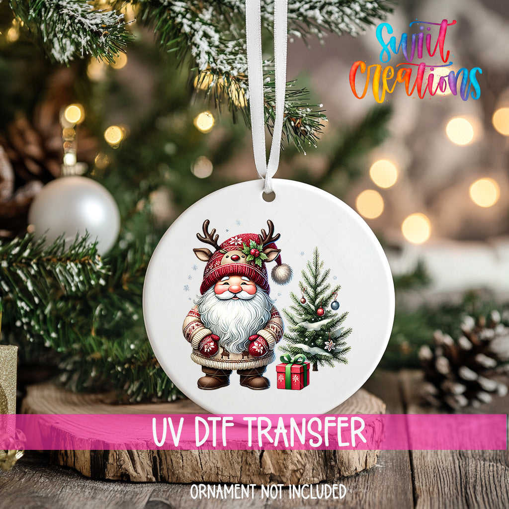 A white ornament with a Santa Claus figure and a Christmas tree is hanging on a tree branch, with a pink ribbon around it.