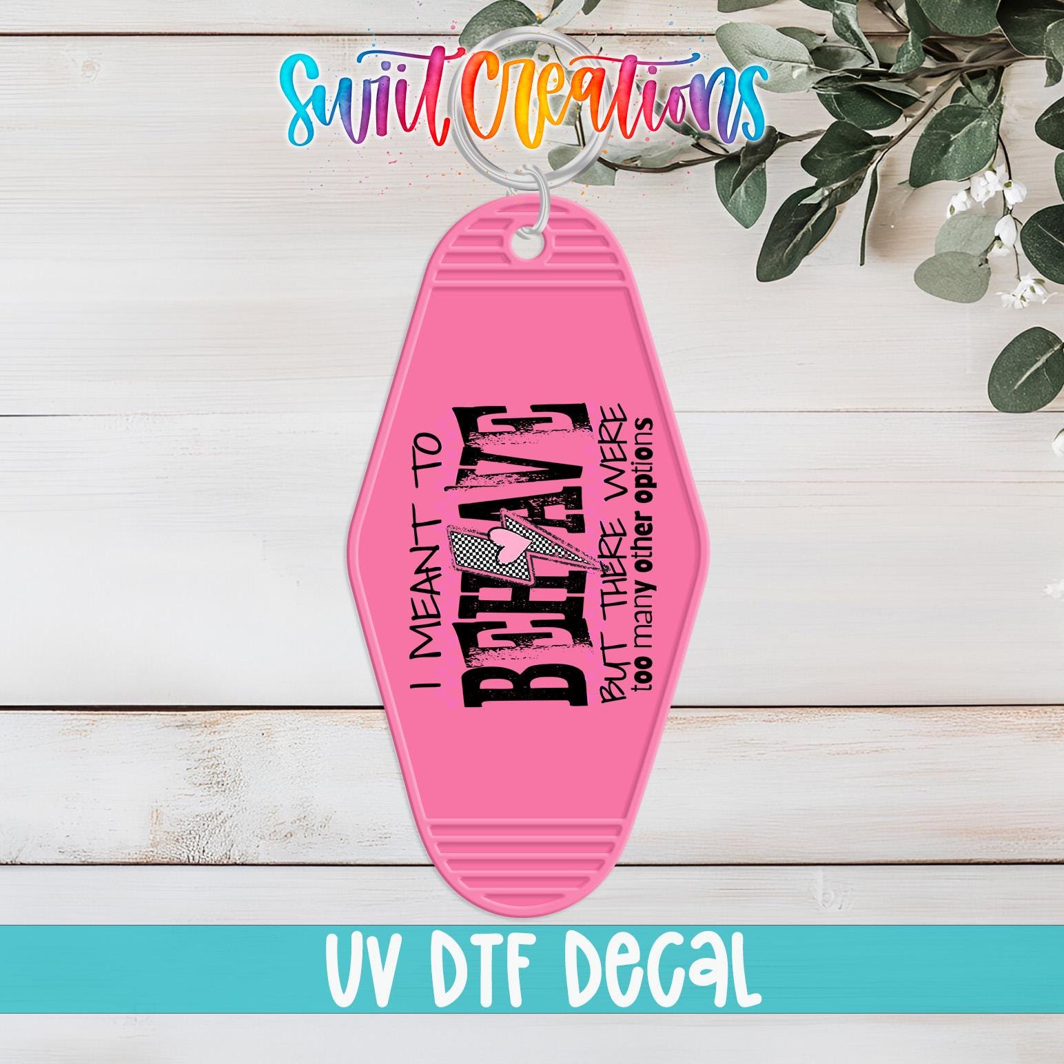 A pink keychain with the text "I MEANT TO BEHAVE BUT THERE ARE THOSE OTHER OPTIONS" printed on it, along with a decorative graphic. The keychain is displayed on a wooden surface, and there is a plant in the background.