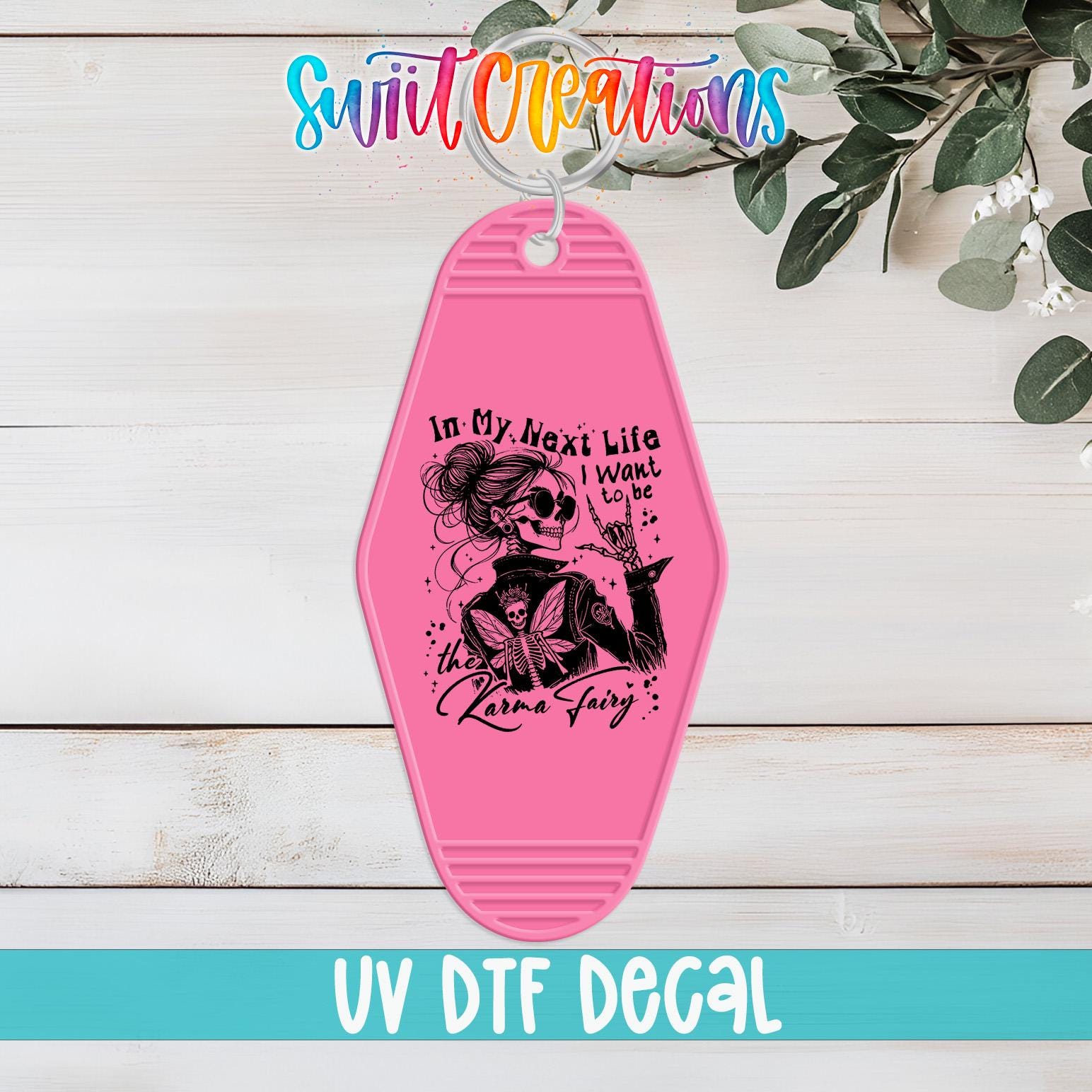 A pink keychain with a skull design and text, placed on a wooden surface with greenery in the background.