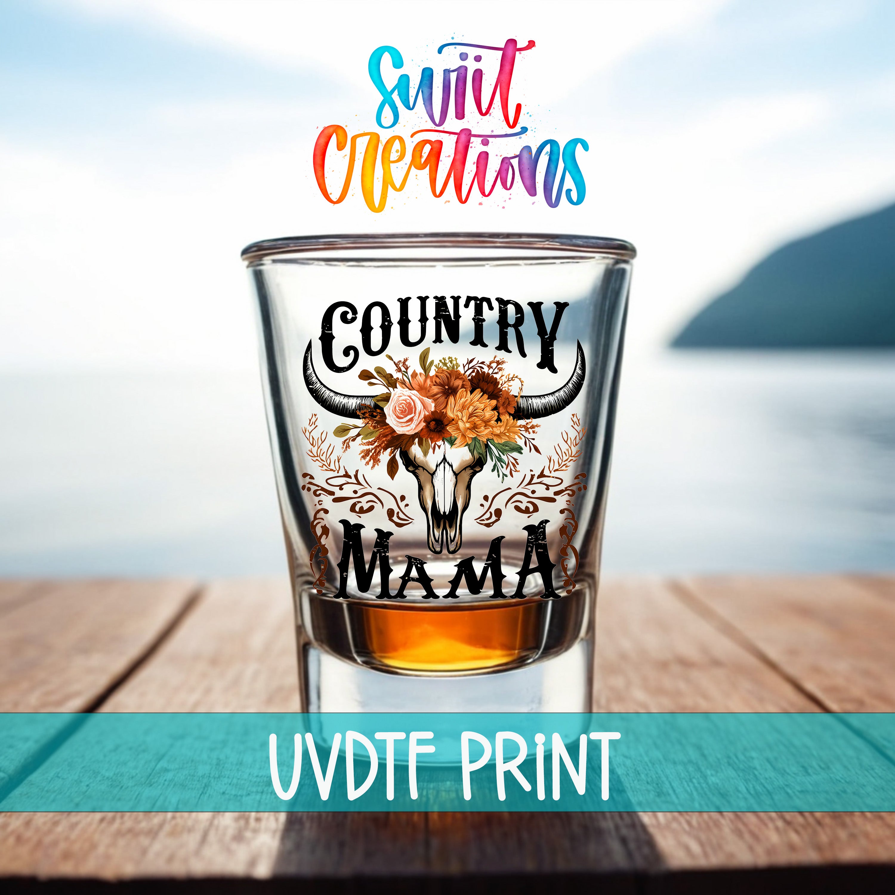 a shot glass with a floral design and the text "COUNTRY MAMA" printed on it, placed on a wooden surface with a blurred background that includes a body of water and a mountain.
