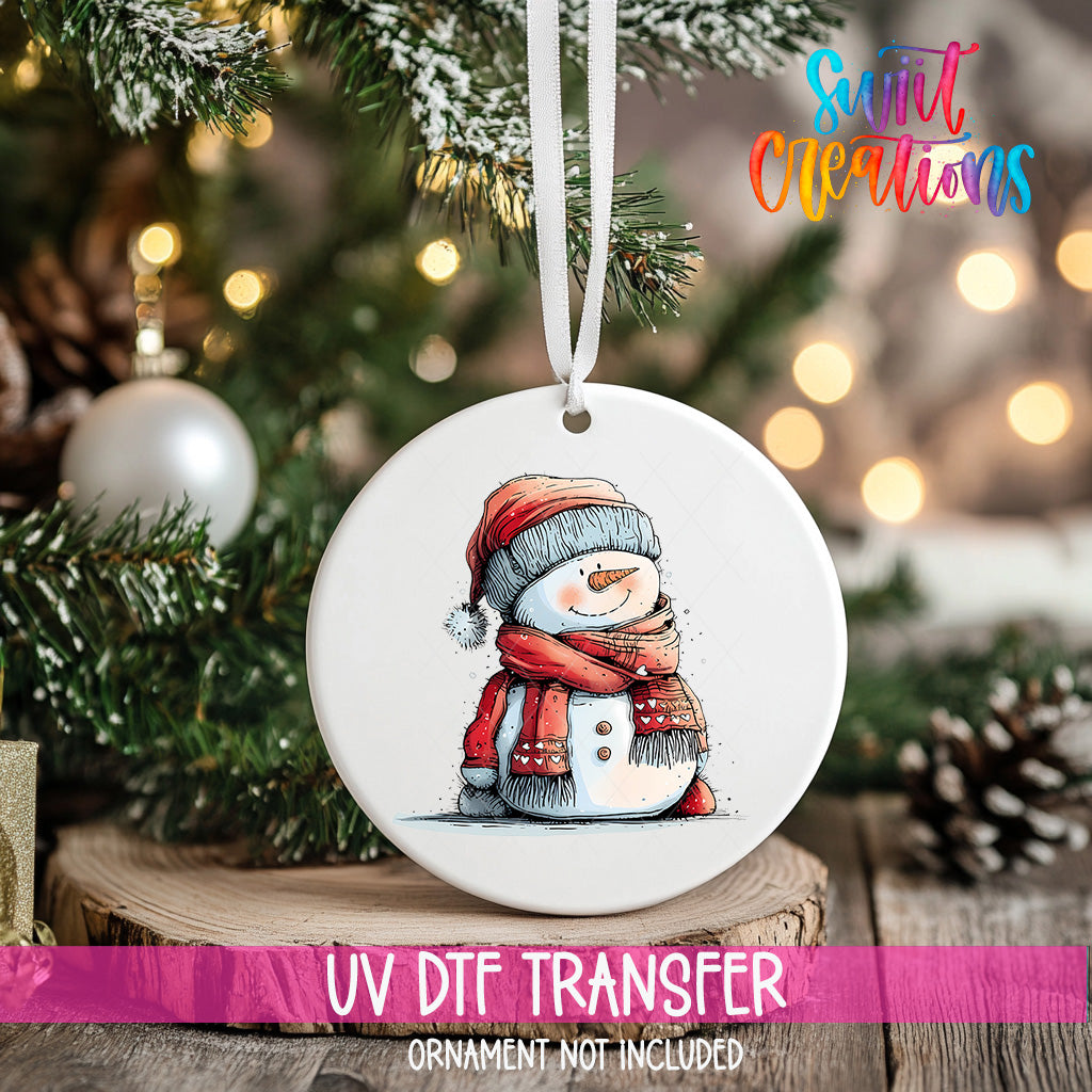 A white ornament with a snowman design hangs on a Christmas tree, surrounded by pine branches and twinkling lights.