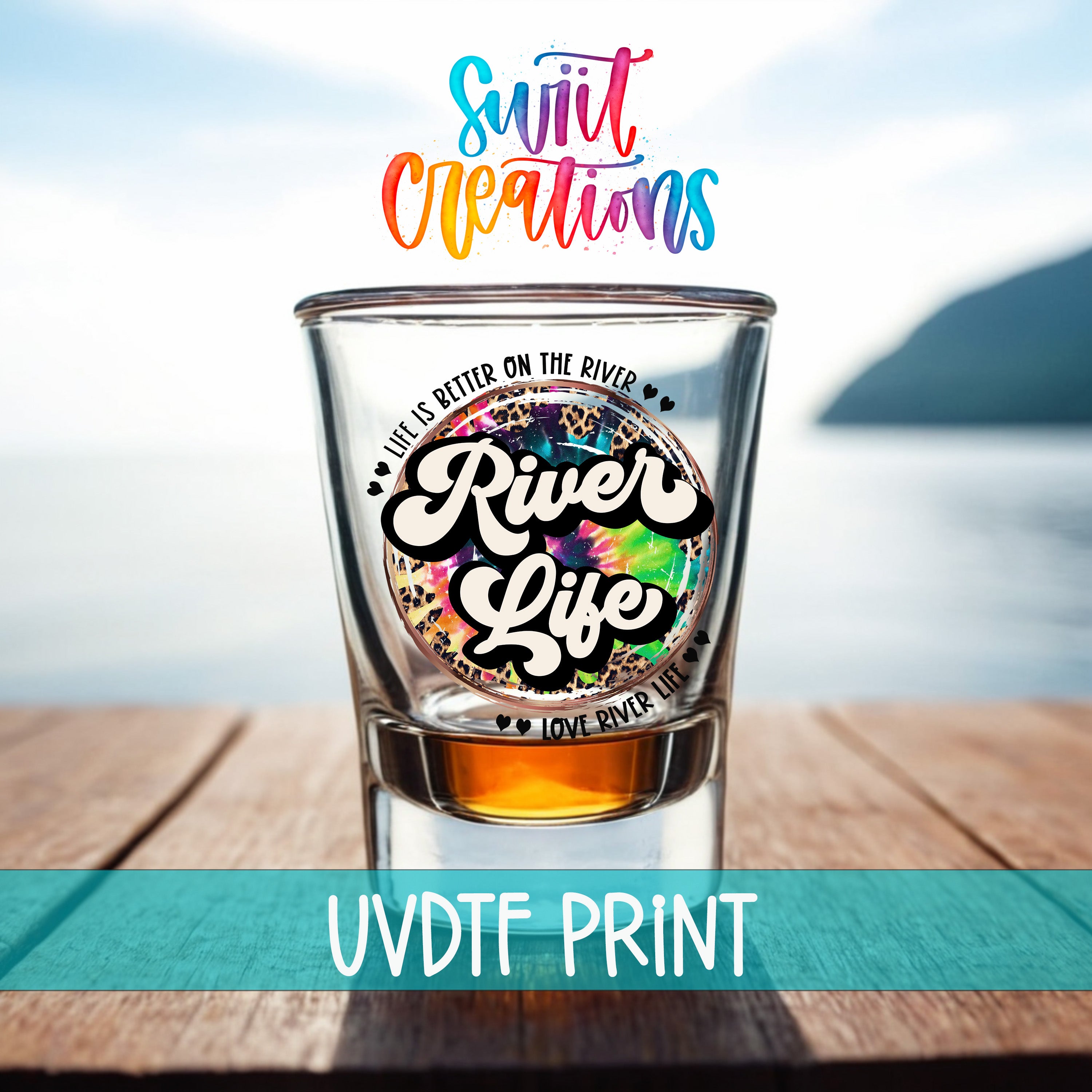 a shot glass with a colorful, tie-dye design and text printed on it, placed on a wooden surface with a body of water in the background.