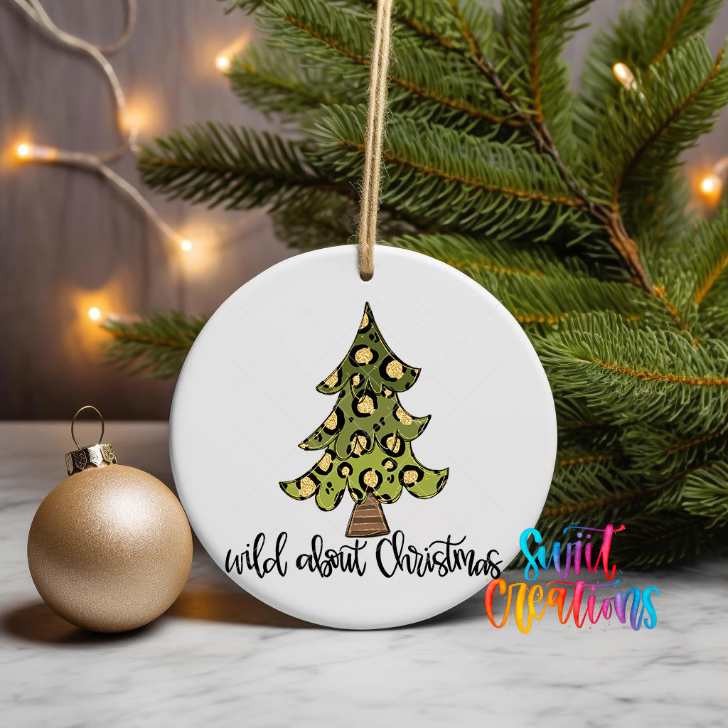 A white Christmas ornament with a leopard print design and the text "wild about Christmas" is displayed on a table, with a gold ornament and a pine tree branch in the background.