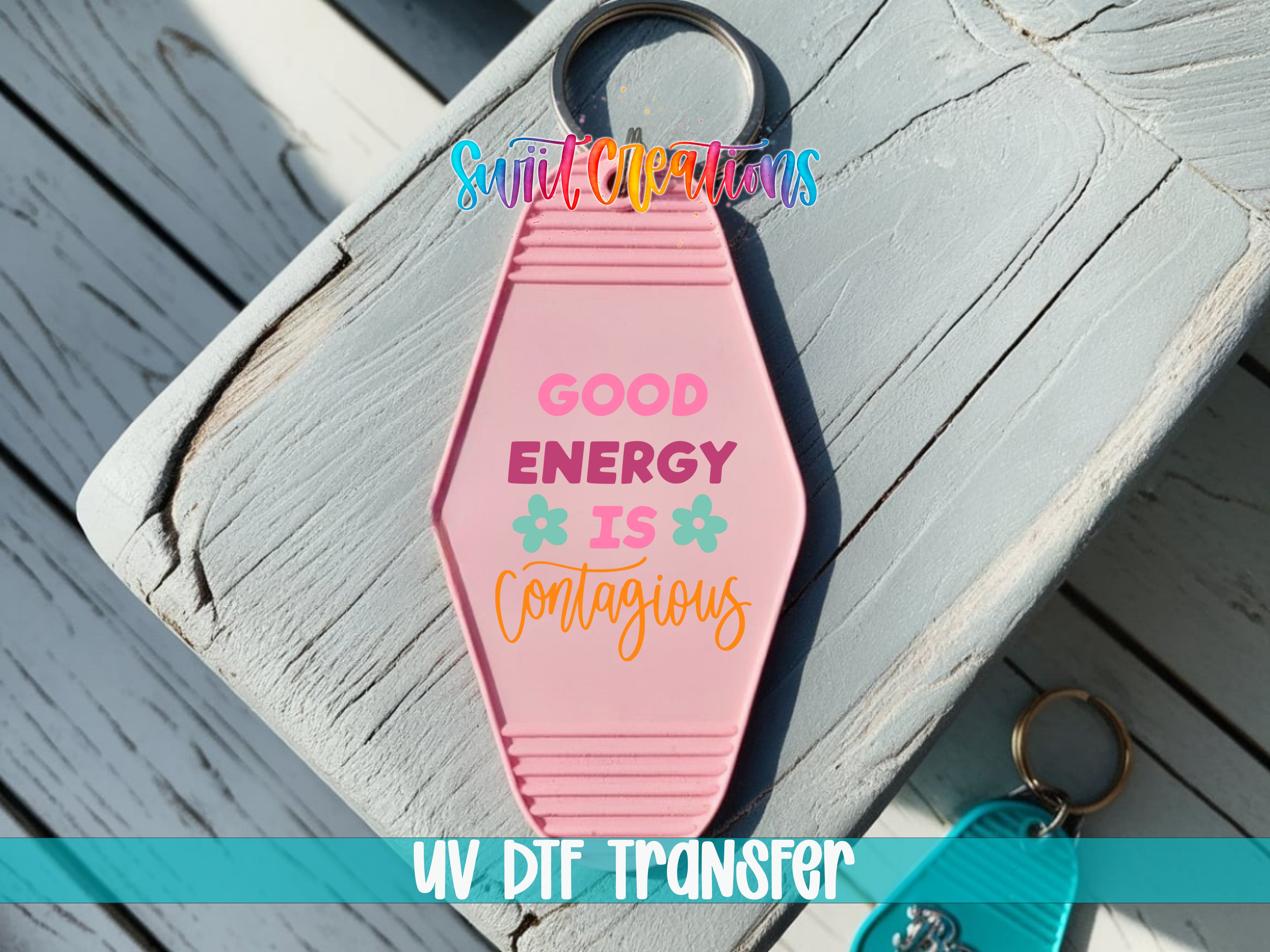 A pink keychain with the text "GOOD ENERGY IS CONTAGIOUS" printed on it, placed on a wooden surface.