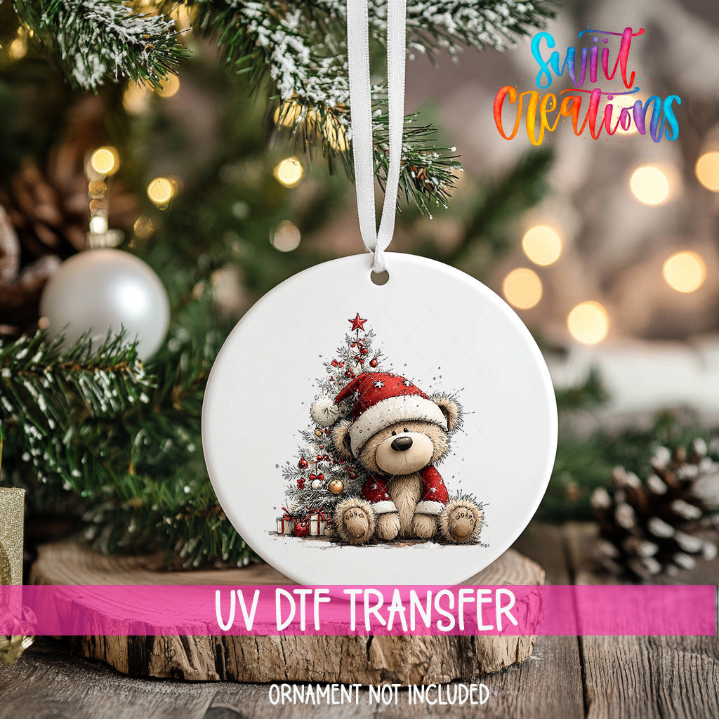 A white Christmas ornament with a teddy bear wearing a Santa hat, sitting next to a decorated Christmas tree, is hanging from a ribbon.