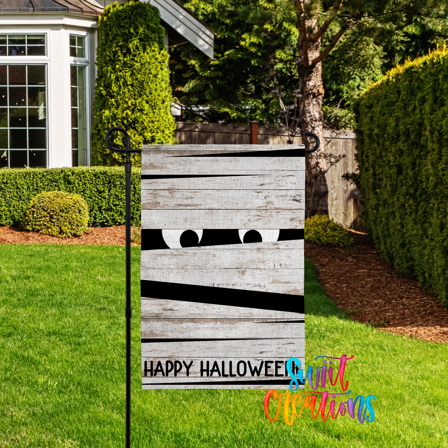 A yard sign with a Halloween-themed design, featuring a pair of eyes peeking out from behind a wooden plank. The sign is located in a yard with a well-maintained lawn and a house in the background.