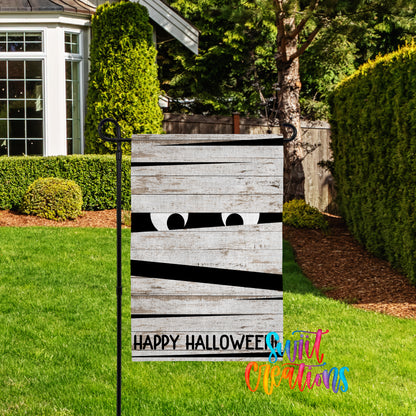 A yard sign with a Halloween-themed design, featuring a pair of eyes peeking out from behind a wooden plank. The sign is located in a yard with a well-maintained lawn and a house in the background.