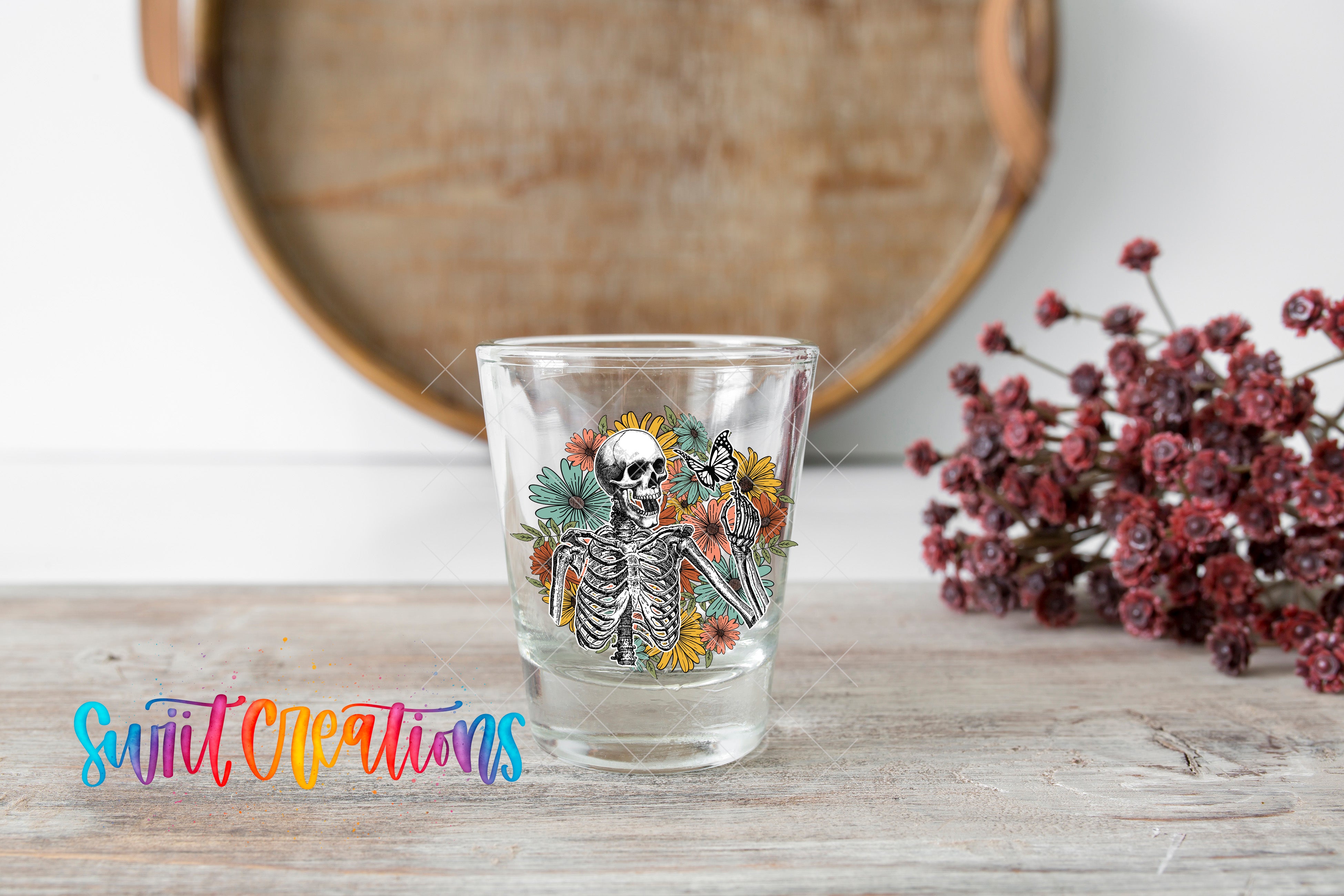 A shot glass with a skeleton design and flowers on a wooden surface, with a wooden tray and red flowers in the background.