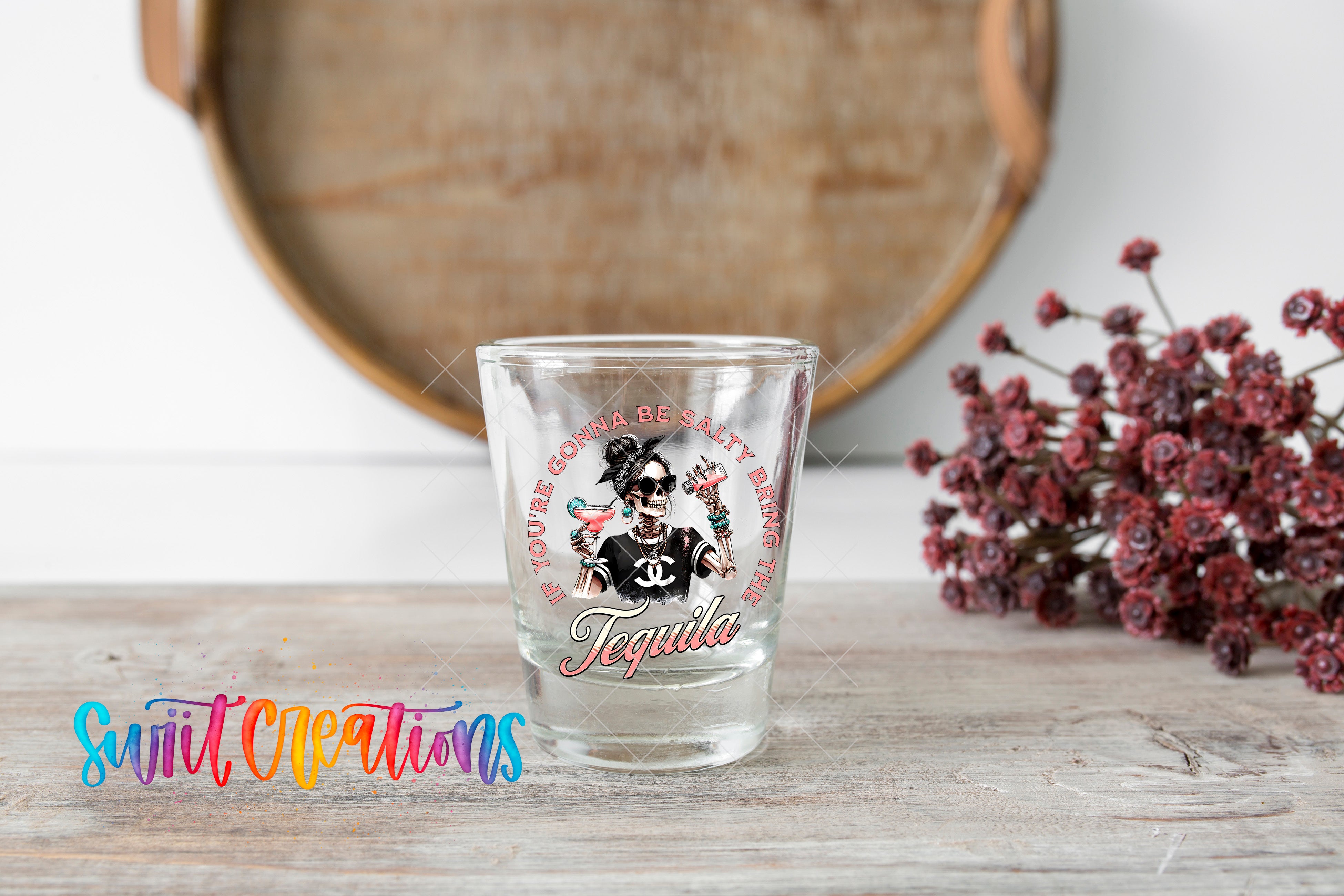 A shot glass with a tequila-themed design and text, placed on a wooden surface next to a bunch of red flowers.