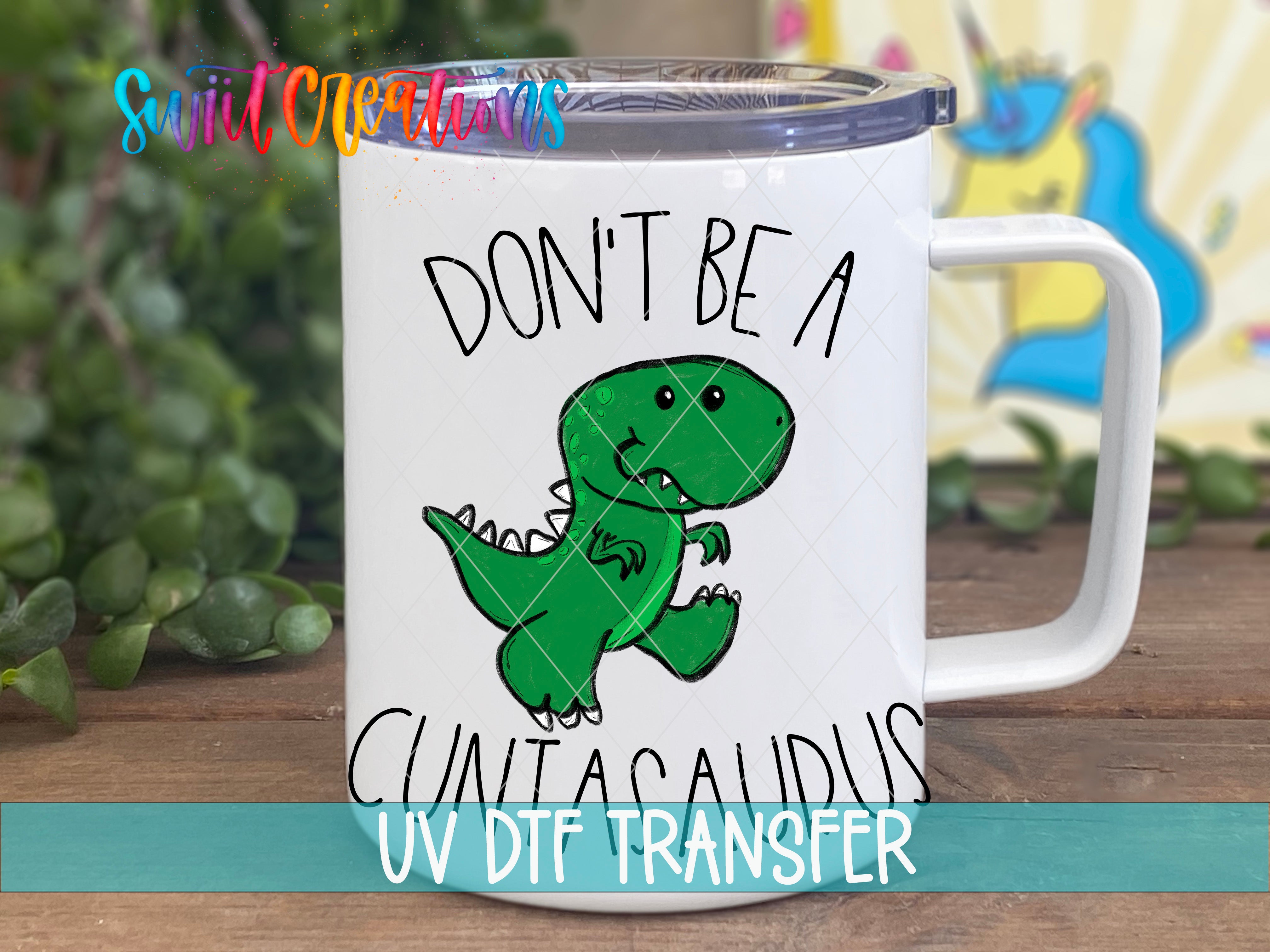 A white mug with a cartoon dinosaur and the text "DON'T BE A CUNTASAUDUS" printed on it.