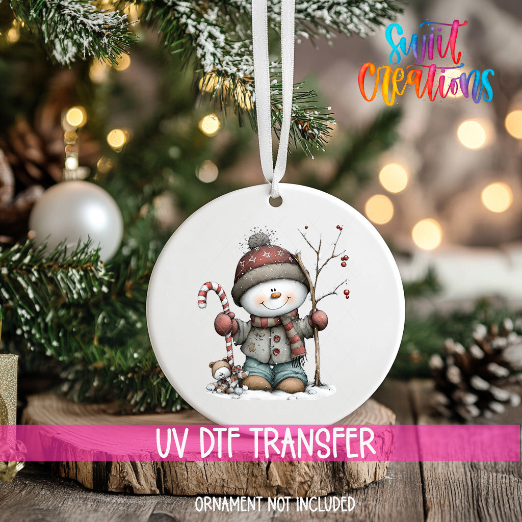 A white ornament with a snowman and candy cane is hanging on a Christmas tree, with a pink ribbon around it.