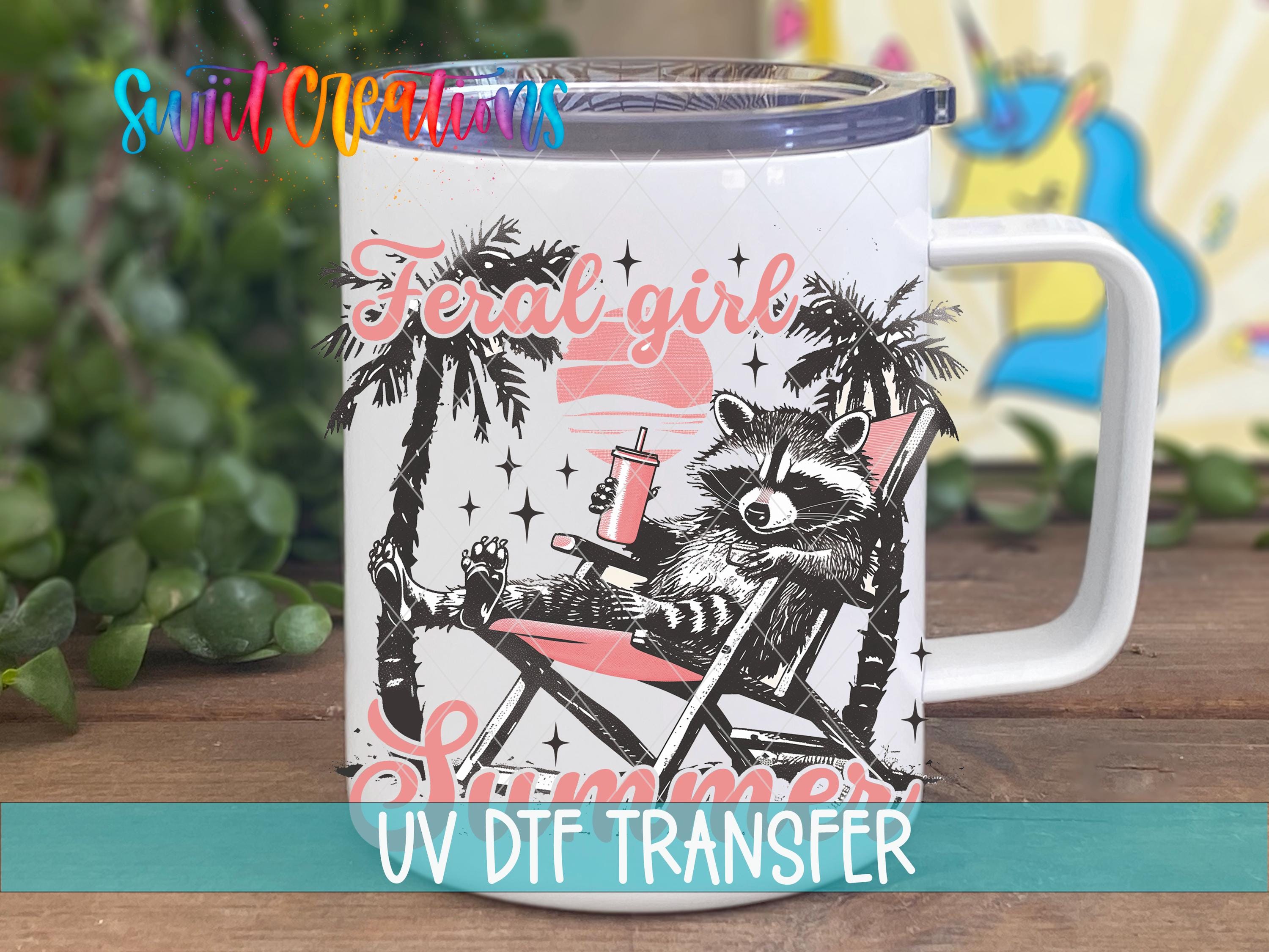 A mug with a raccoon sitting in a beach chair, surrounded by palm trees and a sunset, with the text "Feral Girl Summer" and "UV Dtf Transfer" printed on it.