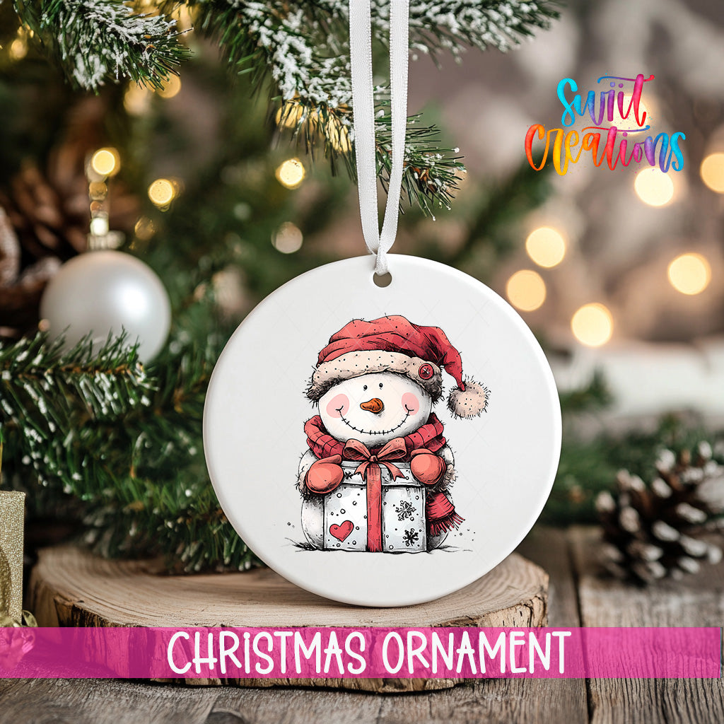 A white Christmas ornament featuring a smiling snowman holding a gift, hanging on a tree branch with a ribbon.