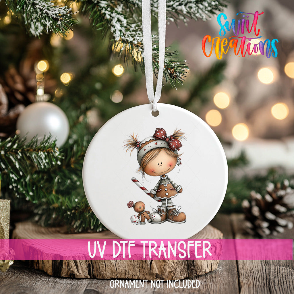 A white ornament with a smiling girl and a gingerbread man is hanging on a Christmas tree, with a pink ribbon around it.
