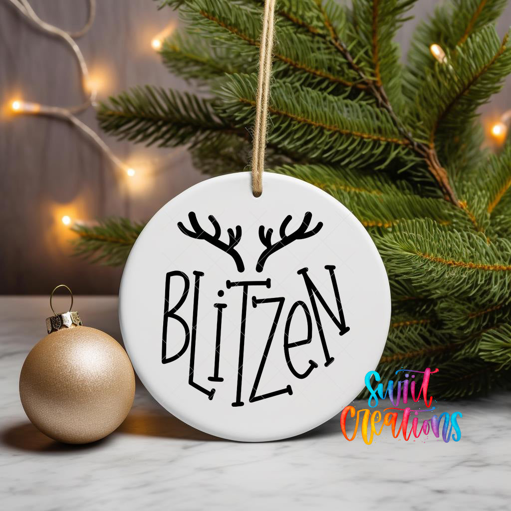 A white ornament with the word "Blitzen" written on it, hanging from a string, is placed next to a gold ornament and a Christmas tree.