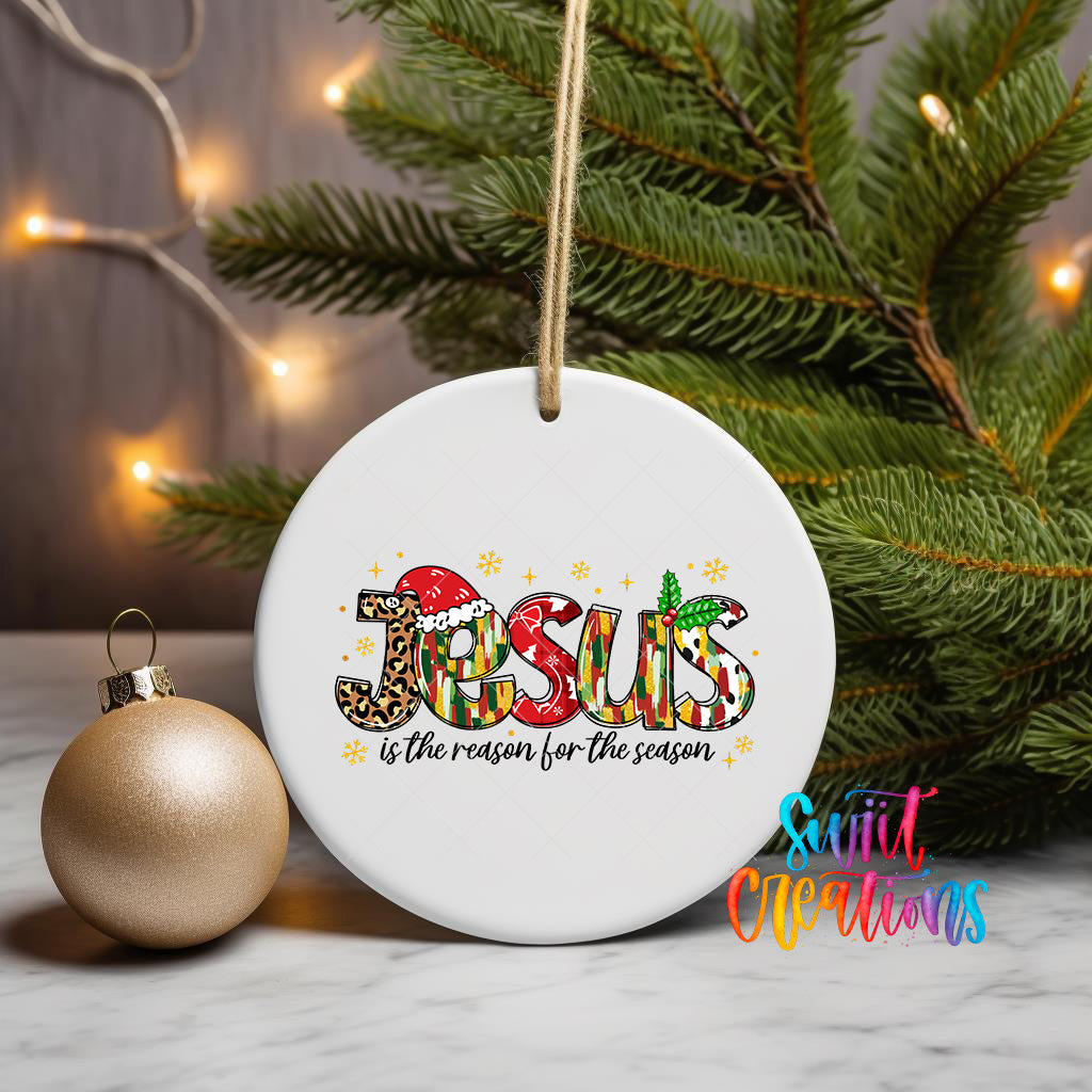 A white ceramic ornament with the word "JESUS" written on it, surrounded by Christmas decorations such as a gold ornament and a pine tree.