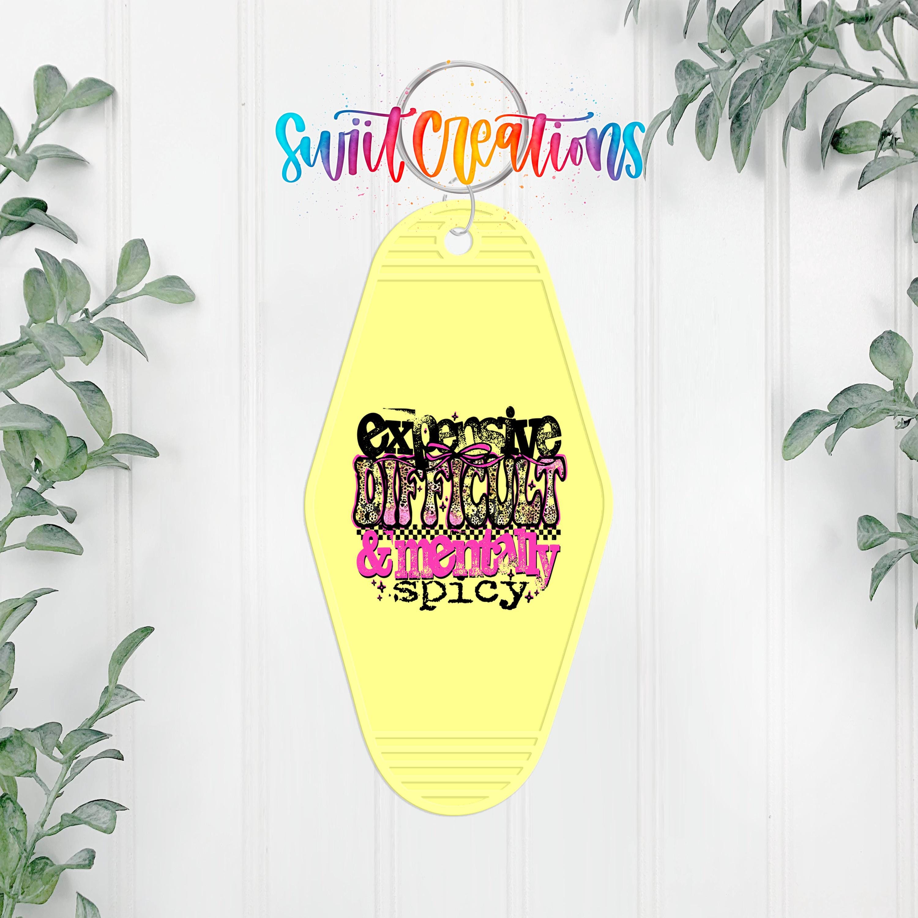 A yellow keychain with the text "Exaggerate Difficulty & Messiness Spicy" printed on it, hanging on a white wall with greenery in the background.
