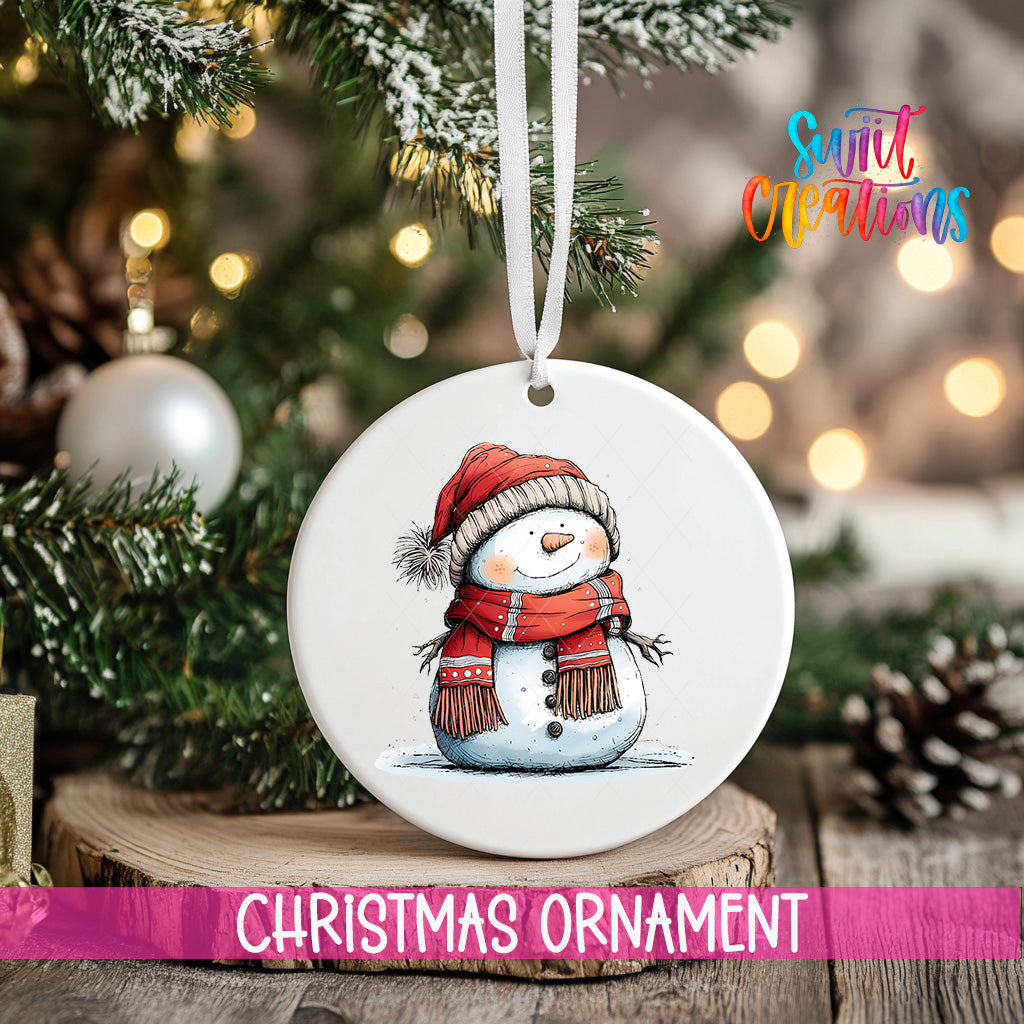 A white Christmas ornament featuring a snowman is hanging from a ribbon, with a blurred background of a decorated Christmas tree and other ornaments.