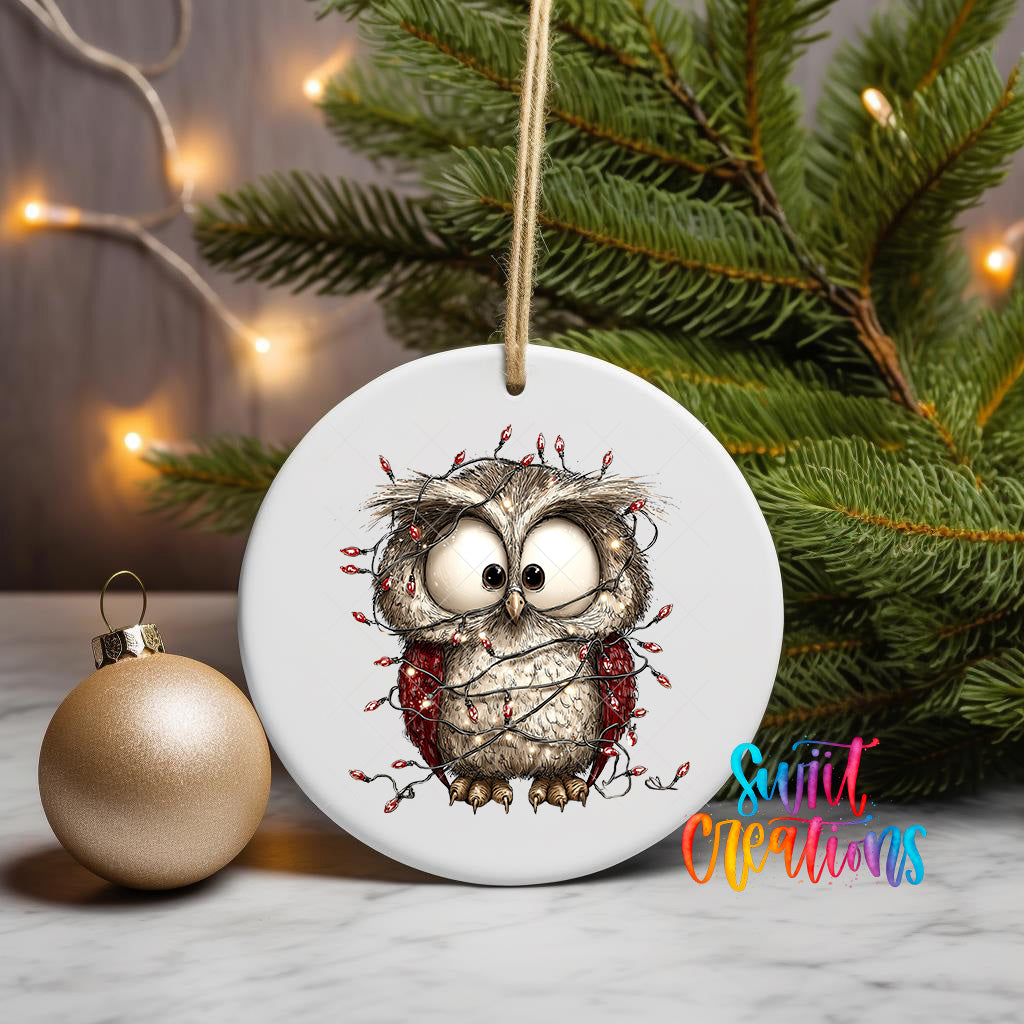 A white ceramic ornament featuring a cartoon owl is displayed on a table, with a gold ornament and a Christmas tree in the background.