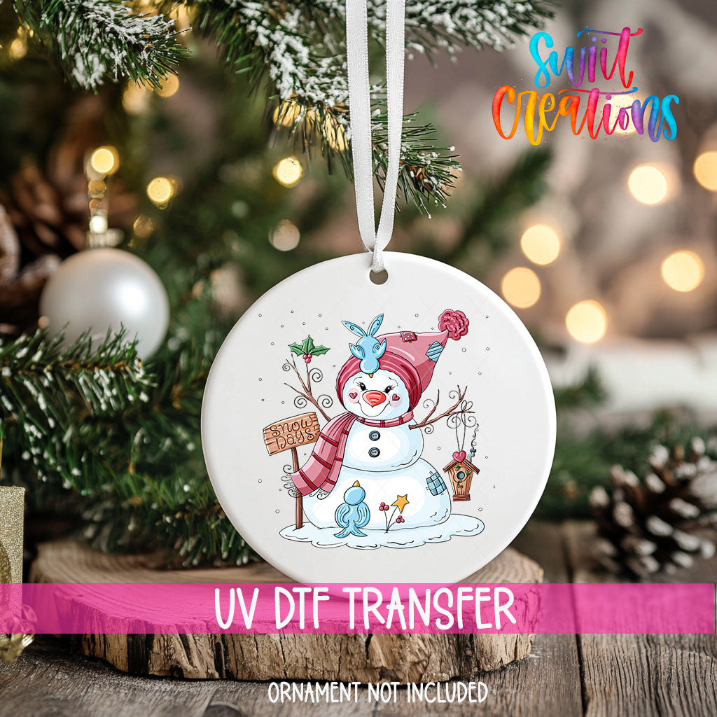 A white ornament with a snowman and other winter-themed elements hangs on a Christmas tree.