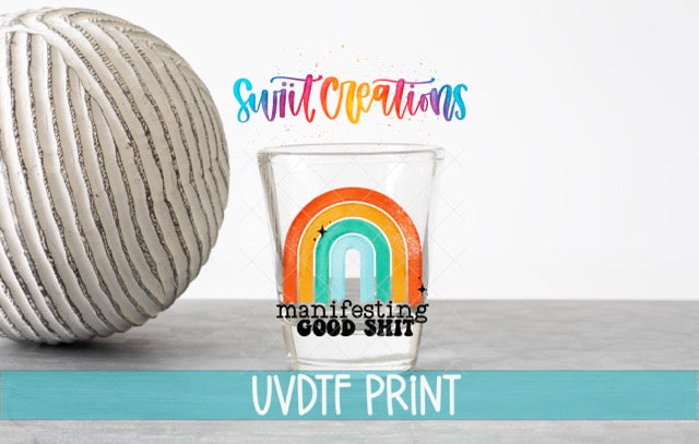 a glass with a rainbow design and the text "manifesting good shit" printed on it, along with a white ball and the text "Swift Creations" and "UVDF PRINT" in the background.