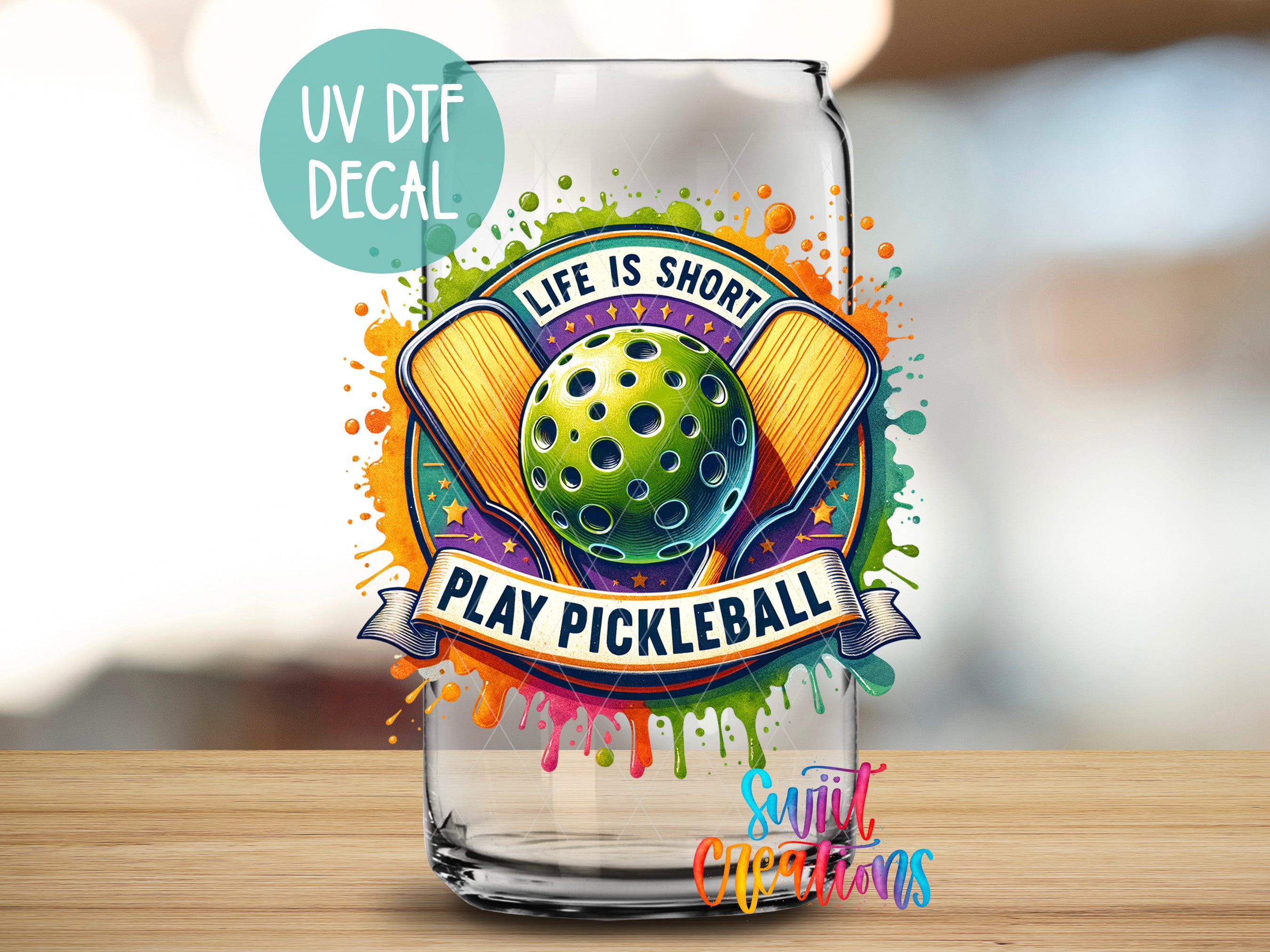a glass can with a colorful and vibrant design, including a tennis racket and a tennis ball, along with the text "Life is short, play pickleball" and "Sunlit Creations".