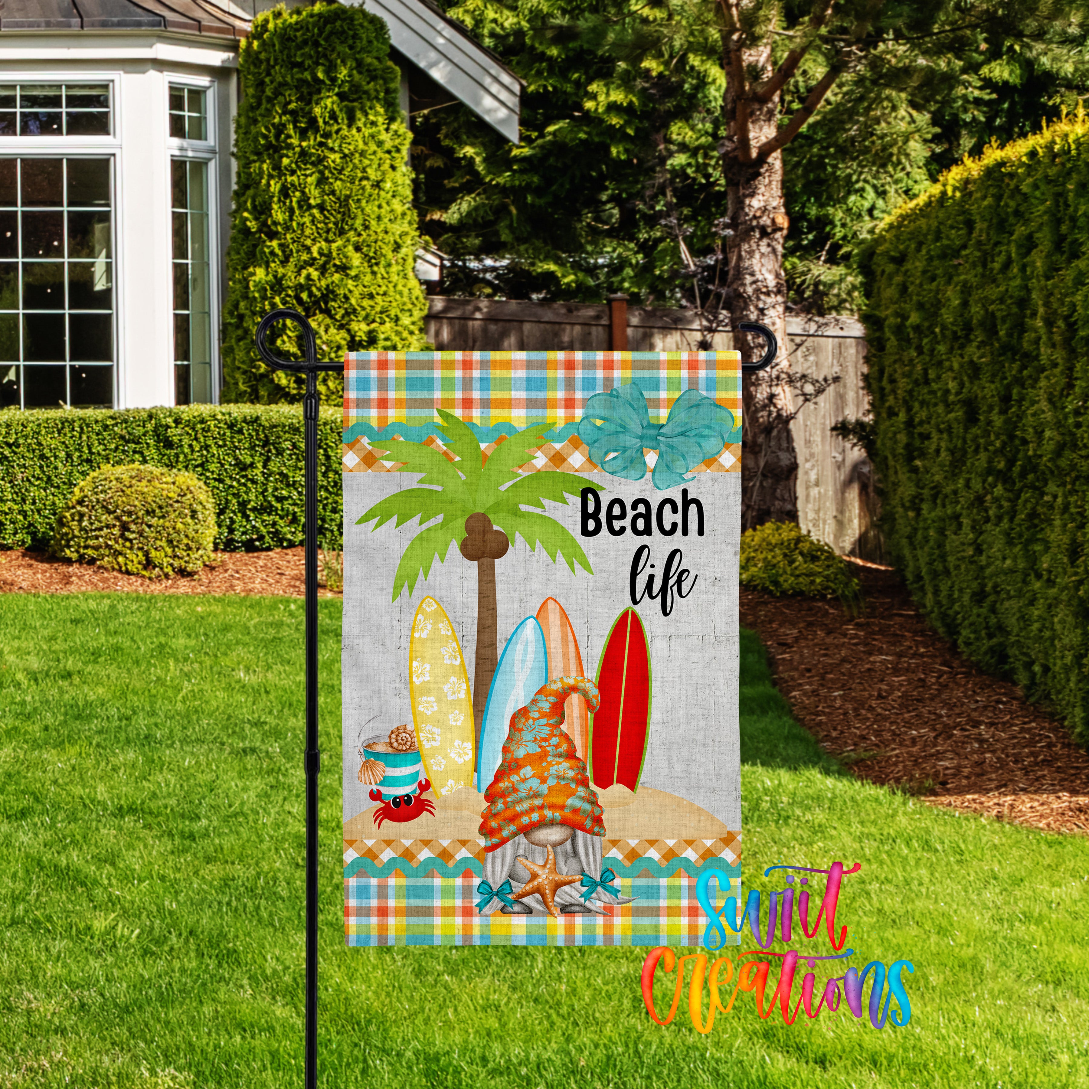 A colorful beach-themed flag is displayed in a grassy yard, featuring a palm tree, surfboards, and a crab.