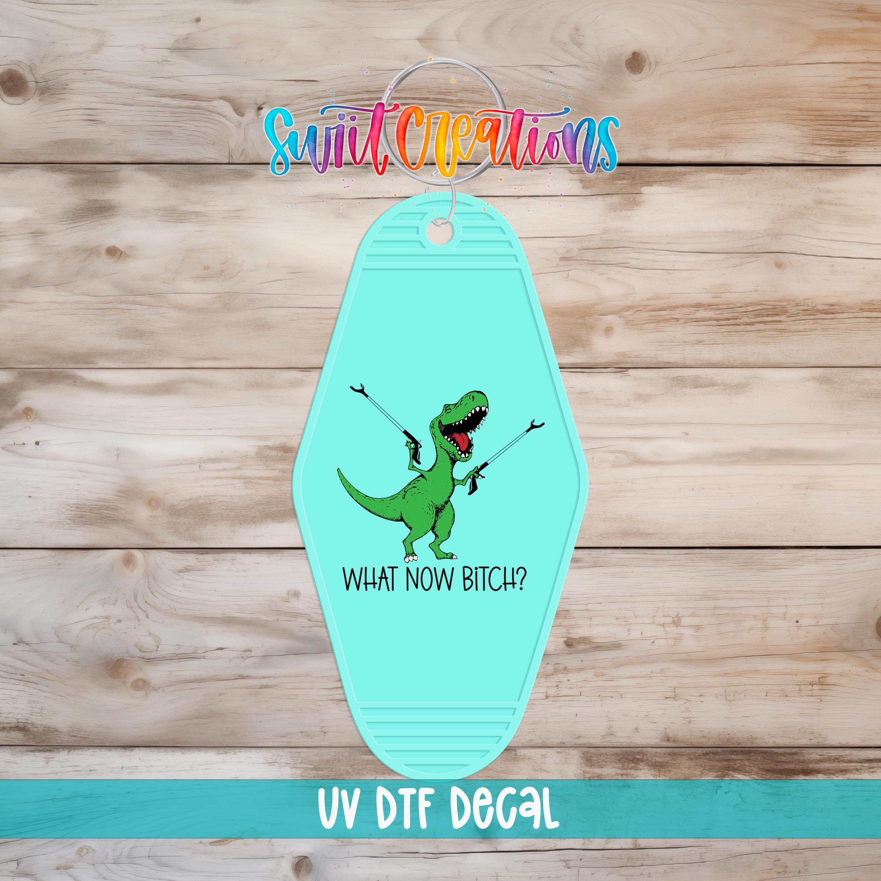 A keychain with a cartoon dinosaur and the text "WHAT NOW BITCH?" printed on it, along with the text "UV Dtf Decal" at the bottom.