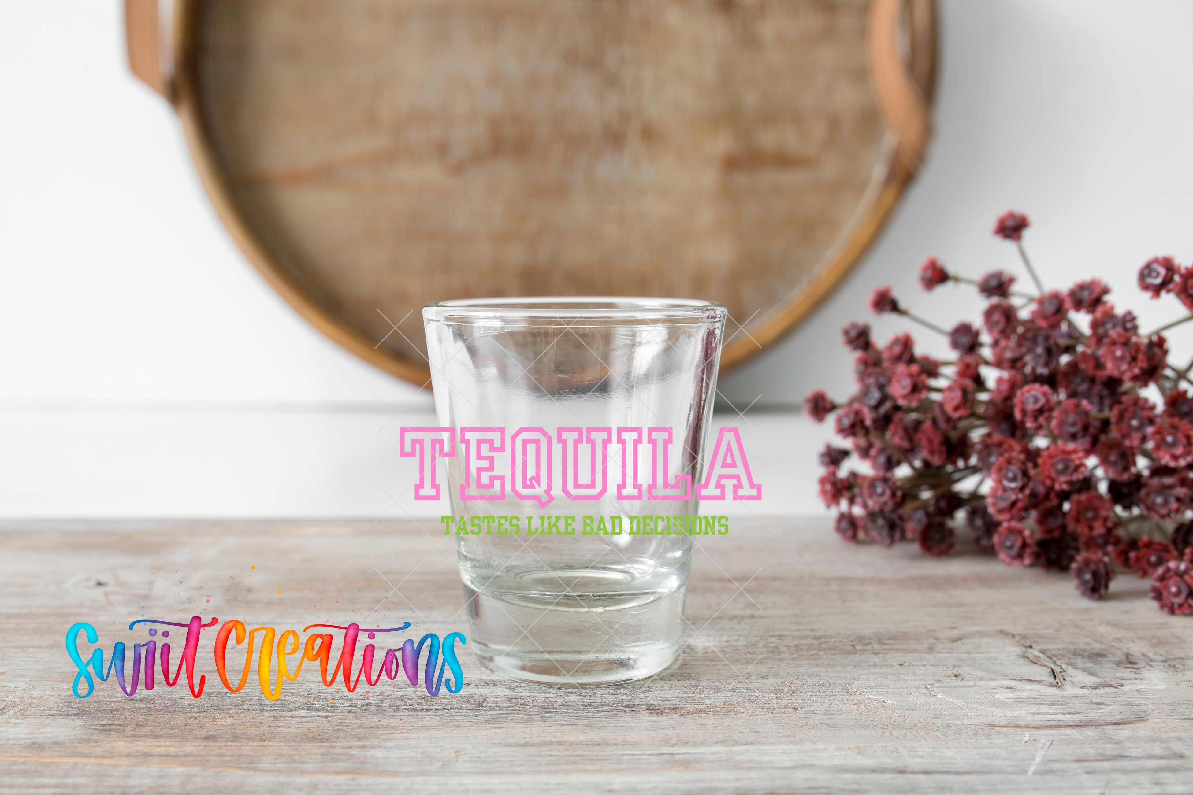 a clear shot glass on a wooden surface, with a bunch of red flowers and a wooden tray in the background.