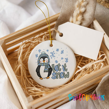 A white ceramic ornament with a penguin design and the text "Let it snow" is displayed in a wooden crate.