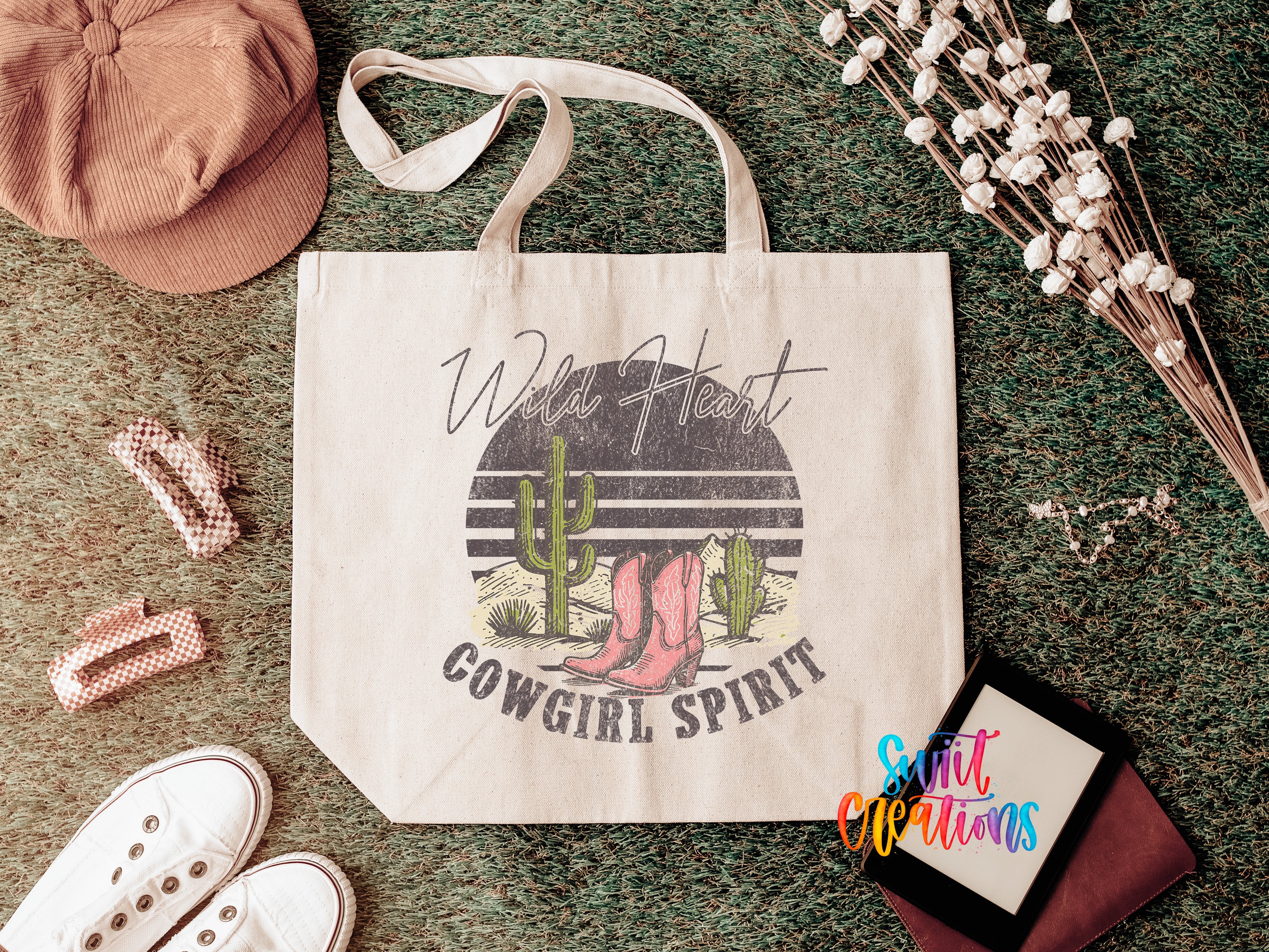 a white tote bag with a graphic design of a cactus and cowboy boots, along with the text "Wild Heart Cowgirl Spirit". The bag is placed on a green carpet, surrounded by various accessories such as a hat, a pair of shoes, and some flowers.