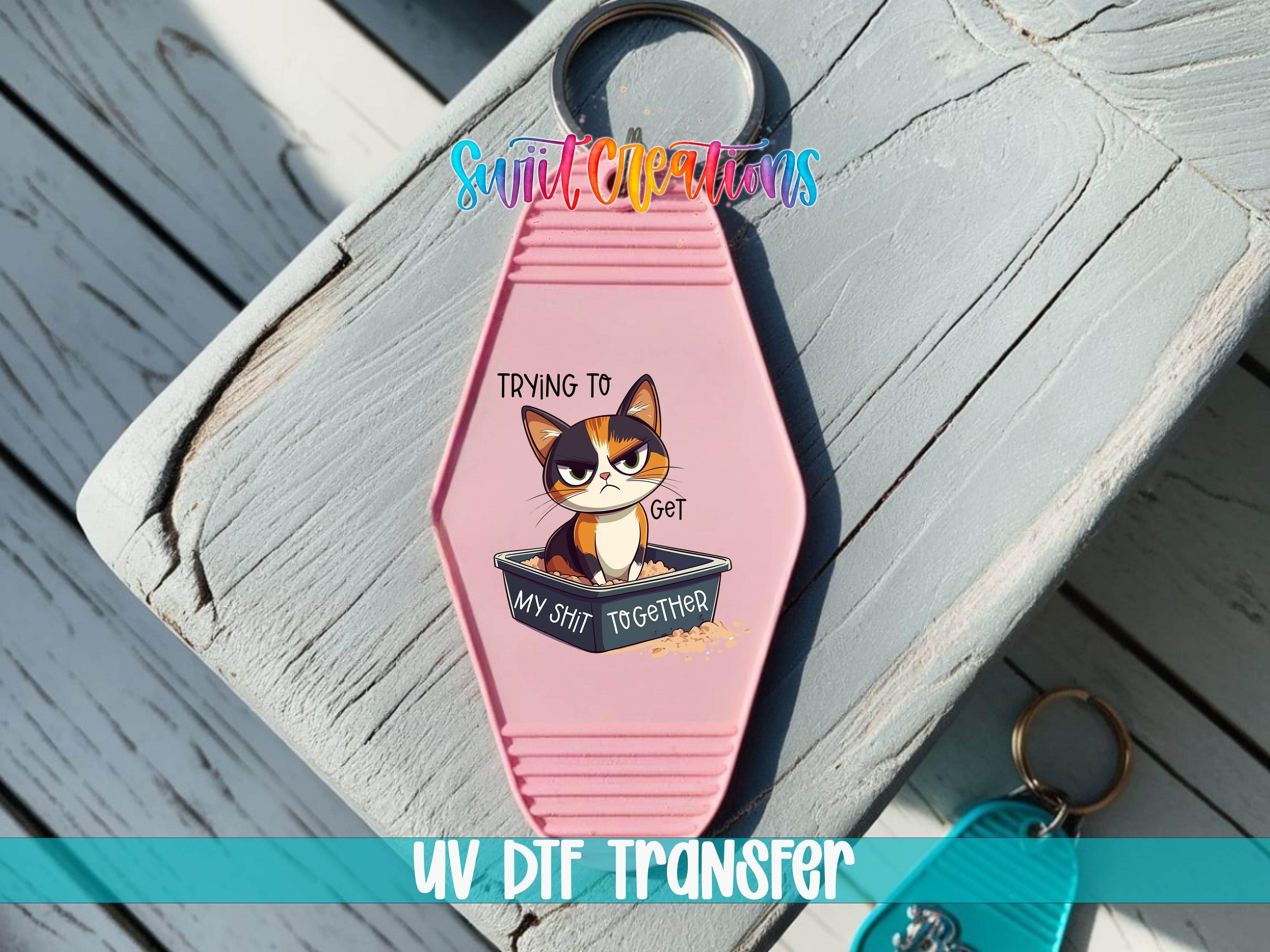 A pink keychain with a cartoon cat and the text "Trying to get my butt together" printed on it, placed on a wooden surface.