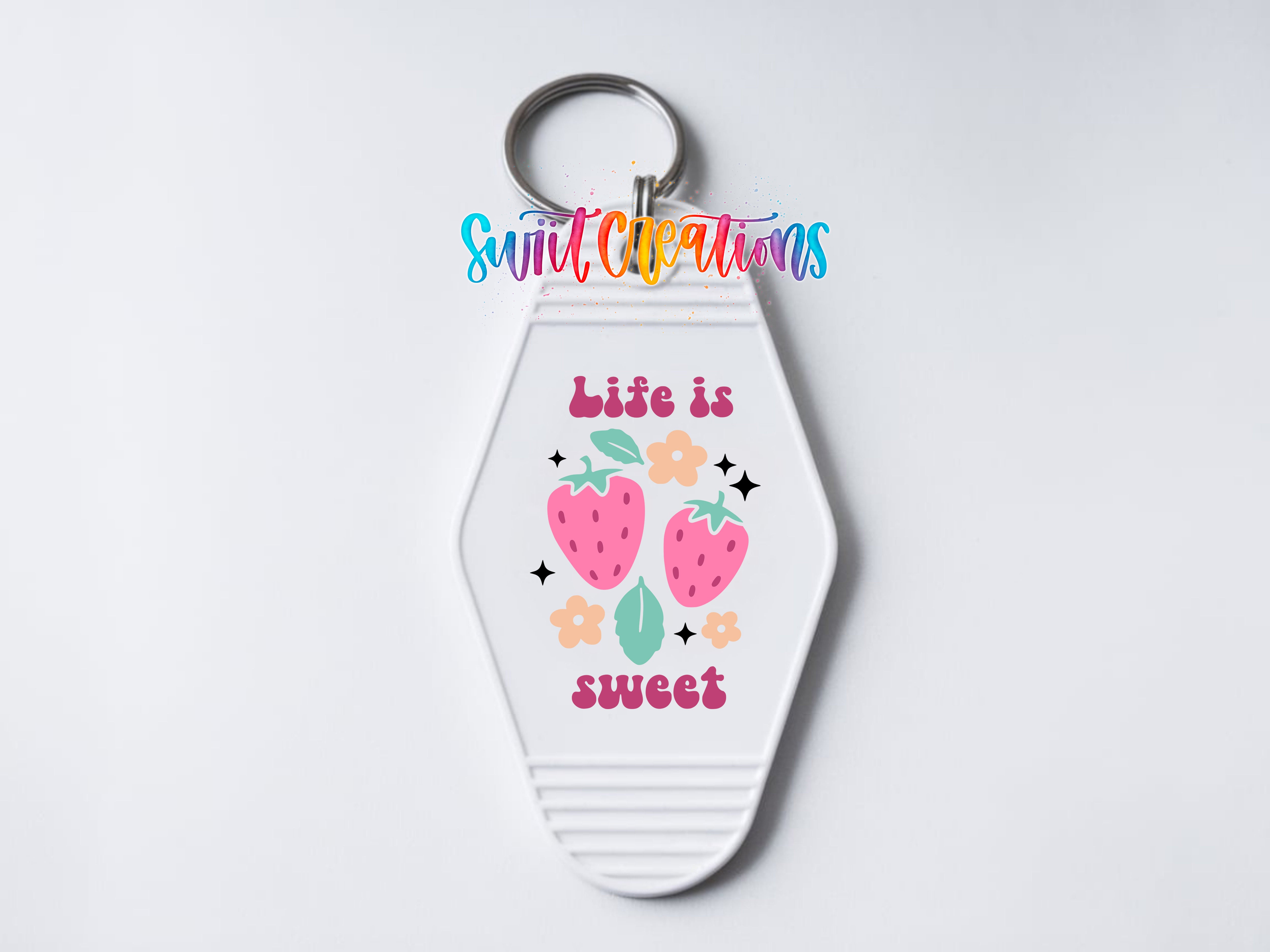 A keychain with a white hexagonal shape and a design featuring two pink strawberries, flowers, and leaves. The text on the keychain reads "Life is sweet".