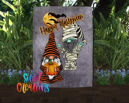 A Halloween-themed garden flag featuring two gnomes, one dressed as a mummy and the other as a gnome, holding pumpkins and surrounded by plants.