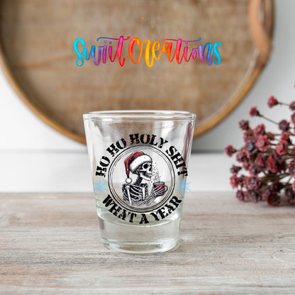 Ho Ho Holy Sh*t What a Year shot glass transfer on a clear glass beside a wooden barrel and dried flowers