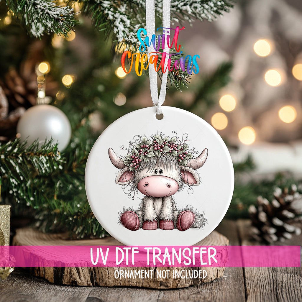 Holly Berry Highland Cow ornament UV DTF transfer on an ornament resting on a wood slice beside a Christmas tree