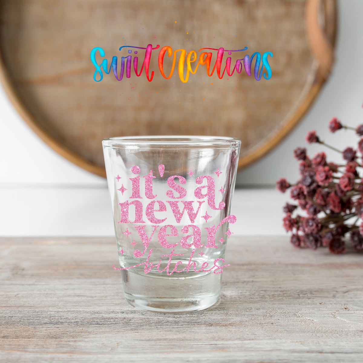 It's A New Year B*tches shot glass transfer on a clear glass beside a wooden barrel and dried flowers