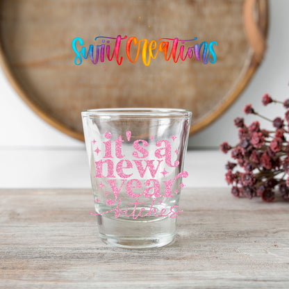 It's A New Year B*tches shot glass transfer on a clear glass beside a wooden barrel and dried flowers