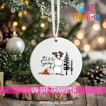Let It Snow Snowman ornament UV DTF transfer on an ornament resting on a wood slice beside a Christmas tree