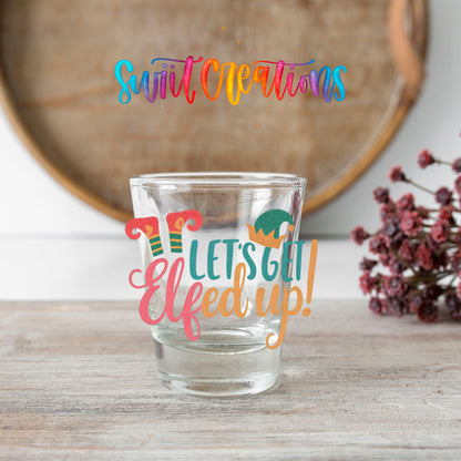Let's Get Elfed Up shot glass transfer on a clear glass beside a wooden barrel and dried flowers