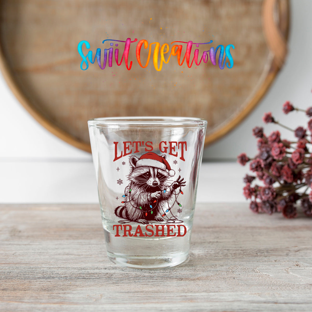 Let's Get Trashed Christmas shot glass transfer on a clear glass beside a wooden barrel and dried flowers
