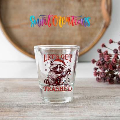 Let's Get Trashed Christmas shot glass transfer on a clear glass beside a wooden barrel and dried flowers