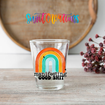 Manifesting Good Sh*t shot glass transfer on a clear glass beside a wooden barrel and dried flowers