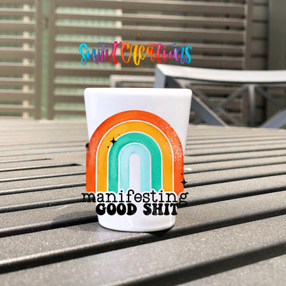 Manifesting Good Sh*t UV DTF transfer on a white shot glass on an outdoor patio table