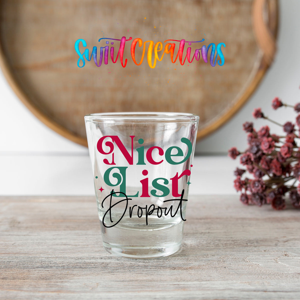 Nice List Dropout Christmas shot glass transfer on a clear glass beside a wooden barrel and dried flowers