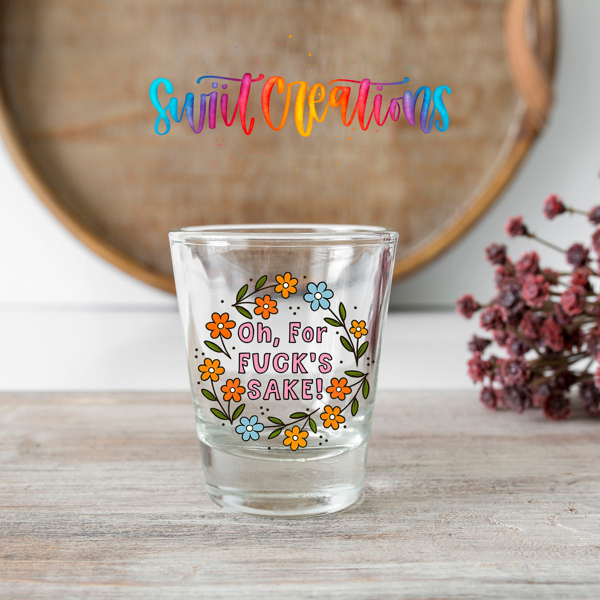 Oh For Fuck's Sake Floral shot glass transfer on a clear glass beside a wooden barrel and dried flowers